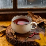 O cană de ceramică albă cu ceai de soc fumegând, într-o atmosferă caldă de toamnă, lângă o fereastră cu picături de ploaie. Ceaiul are o culoare rubinie intensă și strălucitoare. În fundal se văd frunze uscate