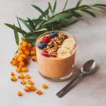 Un smoothie bowl proaspăt. Lângă pahar sunt împrăștiate artistic câteva fructe proaspete de cătină și frunze verzi.