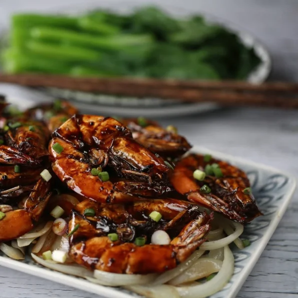 Creveți suculenți glazurați în sos shoyu cu ghimbir, serviți pe un pat de ceapă albă caramelizată și decorați cu ceapă verde proaspătă, prezentați elegant pe o farfurie cu model asiatic.