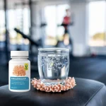 O compoziție care prezintă un pahar cu apă cristalină, cu urme de minerale vizibile, așezat pe un suport cu textură naturală de coral. Fundalul prezintă o zonă de fitness neclară, sugerând energie și vitalitate. Lumina blândă, naturală, subliniază puritatea elementelor.