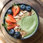 Un smoothie bowl verde proaspăt pregătit, garnisit cu fructe de pădure și semințe, fotografiat de sus pe o masă de lemn deschis la culoare. Smoothie-ul are o textură cremoasă și o nuanță verde vibrantă datorită pulberii de urzică. Lumina naturală cade din lateral, evidențiind prospețimea și textura preparatului.