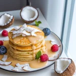 Clătite americane pufoase (pancakes) decorate cu fulgi de cocos proaspăt, zmeură roșie, afine și frunze de mentă, acoperite cu cremă de cocos bio Coconut Bliss Biona, servite pe o farfurie albă cu margine albastră, alături de jumătăți de nucă de cocos proaspătă, într-o prezentare luminoasă lângă fereastră.