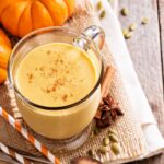 Un smoothie auriu și cremos preparat cu pudră proteică din semințe de dovleac, decorat cu scorțișoară și servit într-o cană de sticlă pe material rustic, alături de dovleac decorativ, semințe de dovleac și anason stelat pentru o aromă de toamnă.