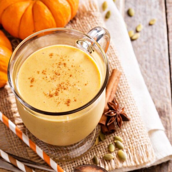 Un smoothie auriu și cremos preparat cu pudră proteică din semințe de dovleac, decorat cu scorțișoară și servit într-o cană de sticlă pe material rustic, alături de dovleac decorativ, semințe de dovleac și anason stelat pentru o aromă de toamnă.