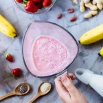 Un smoothie roz cremos cu căpșuni, banane și pulbere de baobab, fotografiat artistic într-un pahar de sticlă, alături de ingredientele sale proaspete precum căpșuni, banane, arahide și linguri din lemn cu semințe și superfood-uri pentru un plus de nutriție.