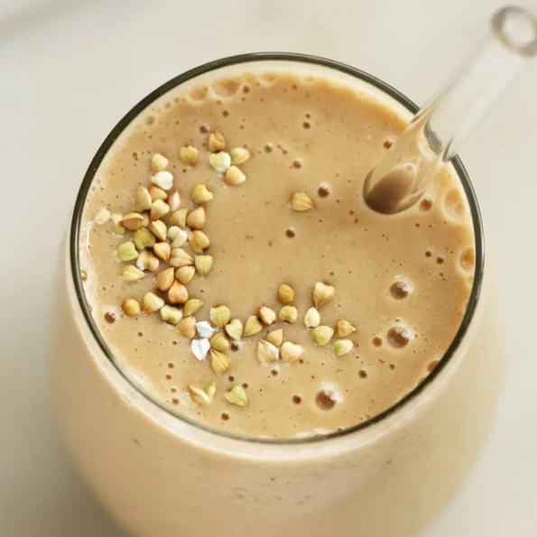 Un smoothie cremos de culoare caramel preparat cu pudră de maca neagră, decorat cu boabe de hrișcă crocantă, servit într-un pahar înalt cu pai, perfect pentru un boost energetic natural.