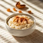 O imagine ce prezintă un bol elegant de ceramică albă plin cu orez cu lapte cremos, garnisit artistic cu scorțișoară pudră. În fundal se observă câteva migdale prăjite și stafide aurii împrăștiate estetic pe o față de masă texturată de culoare crem. Iluminarea naturală vine dintr-o parte, creând umbre fine care scot în evidență textura cremoasă a desertului.