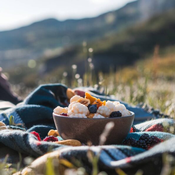 Gustare în natură: În timpul unei pauze de hiking, pe o pătură de picnic în culorile toamnei, este aranjat un bol cu dude albe, alături de alte fructe uscate și nuci. În fundal se vede un peisaj montan blur, iar lumina naturală a după-amiezii creează o atmosferă caldă și reconfortantă.