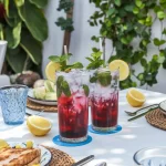 Un aranjament pe o masă de terasă în timpul unui brunch în aer liber. În centru, două pahare înalte cu băutură de culoare roșu-închis diluată cu apă minerală și decorată cu frunze de mentă și felii de lămâie. În fundal se văd elemente de decor mediteraneean și vegetație verde, sugerând un stil de viață sănătos și natural.