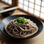 O farfurie ceramică tradițională japoneză neagră cu tăiței Soba aburinzi, garnisiți cu ceapă verde proaspătă tăiată fin și susan negru presărat deasupra. În fundal se văd bețișoare din bambus și un bol mic cu sos tsuyu