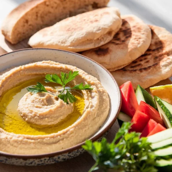 O masă mediteraneană în lumină naturală, cu un bol de hummus proaspăt preparat în prim-plan, decorat artistic cu ulei de măsline și pătrunjel. Lângă bol, câteva lipii calde și legume proaspete tăiate pentru servire. Imaginea transmite prospețime și autenticitate culinară