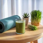 A peaceful morning wellness scene showing a glass of fresh green juice on a wooden table next to a yoga mat. Soft natural lighting highlights the vibrant green color of the drink. Some fresh wheat grass plants in the background create a connection to the natural source. The composition suggests a moment of mindful healthy living.
