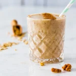 Un smoothie cremos de culoare bej în pahar de cristal cu model în relief, decorat cu scorțișoară pudră și servit cu pai eco, alături de nuci pentru decor, perfect pentru un mic dejun sănătos și energizant.