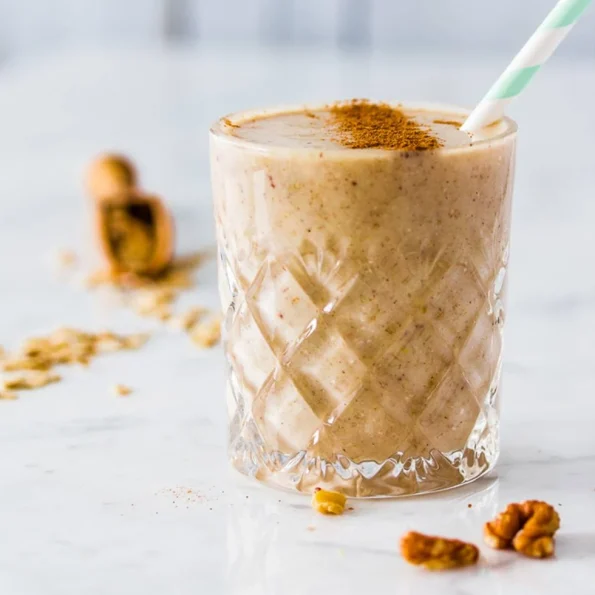 Un smoothie cremos de culoare bej în pahar de cristal cu model în relief, decorat cu scorțișoară pudră și servit cu pai eco, alături de nuci pentru decor, perfect pentru un mic dejun sănătos și energizant.