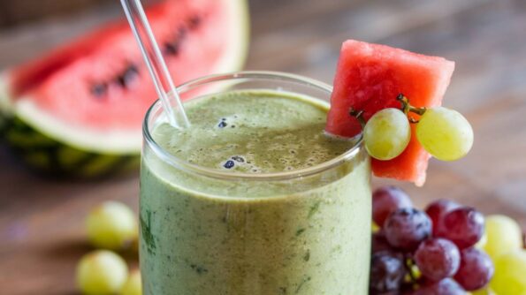 5 Remedii Care Te Ajută Să Reduci Oboseala Musculară Un smoothie verde proaspăt și răcoritor decorat cu pepene roșu și struguri, servit lângă fructe proaspete pe o masă de lemn.